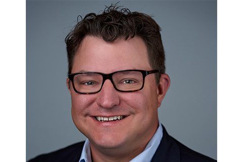 A man with short brown hair and glasses smiles at the camera, wearing a dark jacket over a light shirt, against a plain gray background.