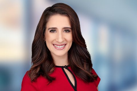 A woman with long brown hair wearing a red top smiles at the camera, standing in front of a blurred indoor background.