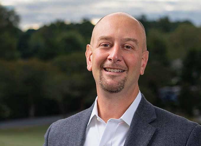 A bald man with a goatee, wearing a white shirt and gray blazer, stands outdoors and smiles at the camera with trees and a cloudy sky in the background.