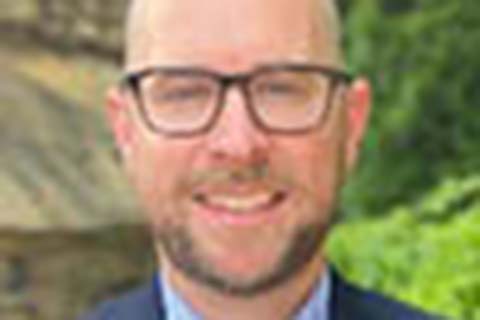 A man with glasses and a beard wearing a suit jacket and blue shirt is standing outdoors with greenery in the background.