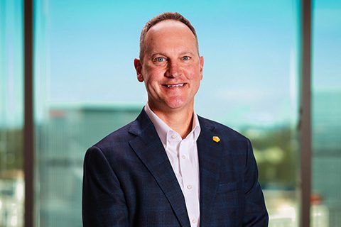 Dark haired man in a blazer smiles in front of a bright window with a blurred city view