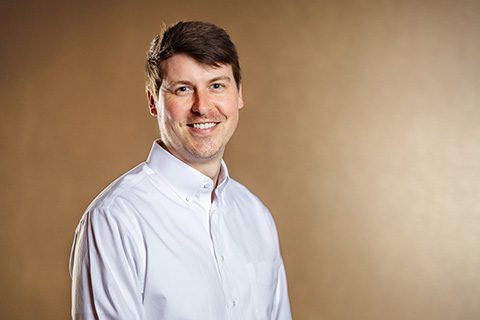 A man wearing a white button-up shirt smiles at the camera against a plain, light brown background.