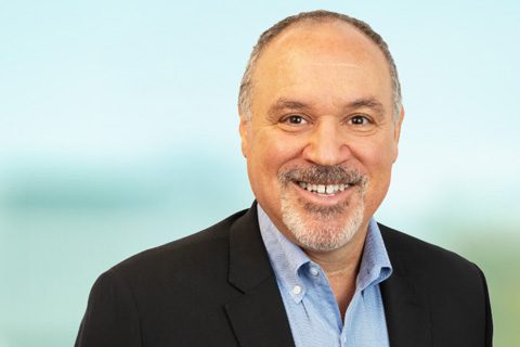 Smiling middle-aged man with short gray hair and beard, wearing a black blazer and light blue shirt, standing against a blurred light blue background.