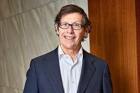 A man in a suit and glasses stands indoors, smiling at the camera, with a marble wall and wooden panel in the background.