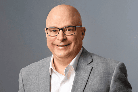 Bald man wearing glasses, a white shirt, and a light gray suit jacket, smiling in front of a plain gray background.