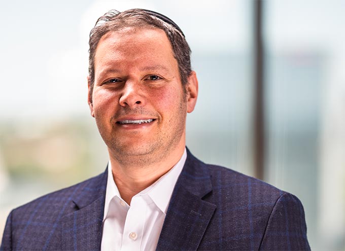 A man in a dark plaid suit jacket and white shirt stands indoors in front of a blurred window background, smiling at the camera.