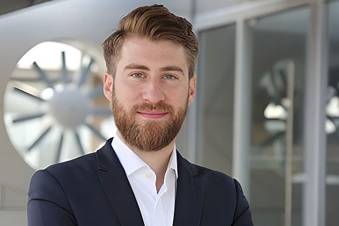 A man with brown hair and a beard wearing a dark suit and white shirt stands in front of a modern building with large windows and a turbine in the background.