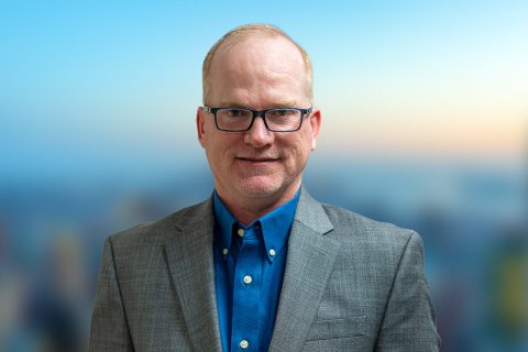 Man in glasses and a gray blazer smiles in front of a bright window with a blurred city view