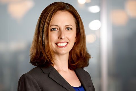 A woman with shoulder-length brown hair, wearing a grey blazer and blue top, smiles at the camera against a blurred office background.