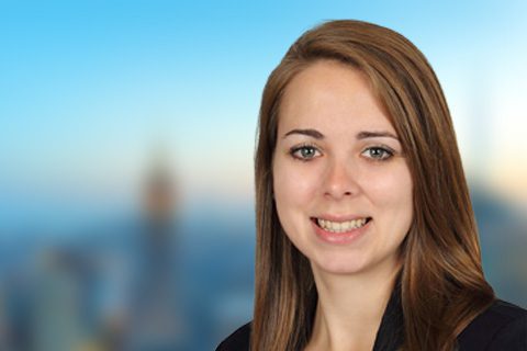 A woman with straight brown hair, wearing a black top, smiles at the camera with a blurred cityscape background.