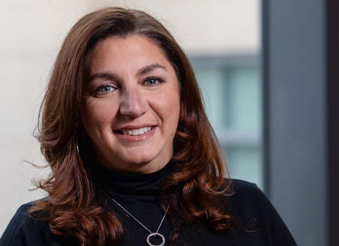 A woman with long brown hair wearing a black turtleneck and a silver necklace, smiling in front of a window and blurred background.