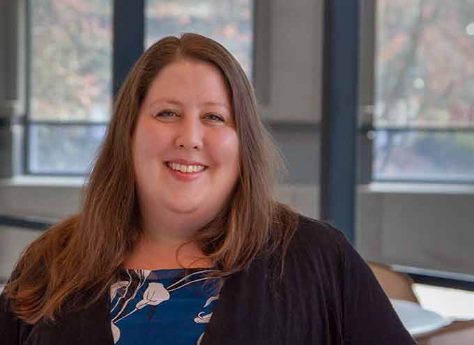 A woman with long brown hair, wearing a dark jacket and patterned top, smiles while standing indoors in front of large windows.
