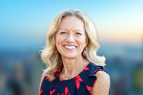 Blonde woman in a navy and red sleeveless top smiles in front of a bright window with a blurred city view.