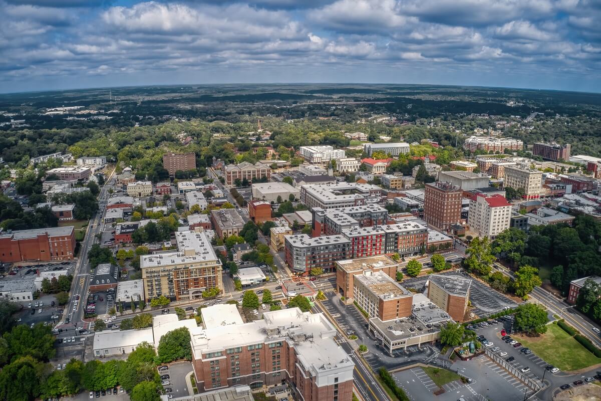 Aerial view of a small city with a mix of commercial, residential, and green areas under a partly cloudy sky.