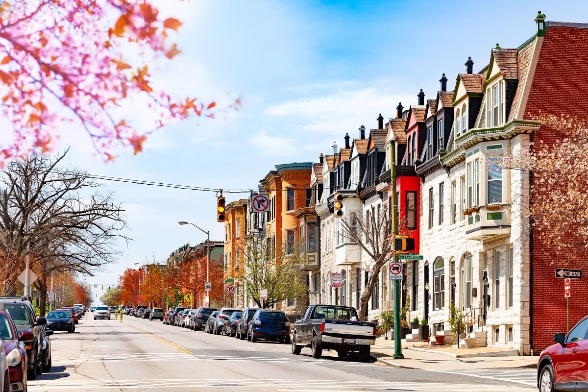 A city street lined with parked cars and colorful, historic row houses on a clear day with light traffic; trees show spring foliage.