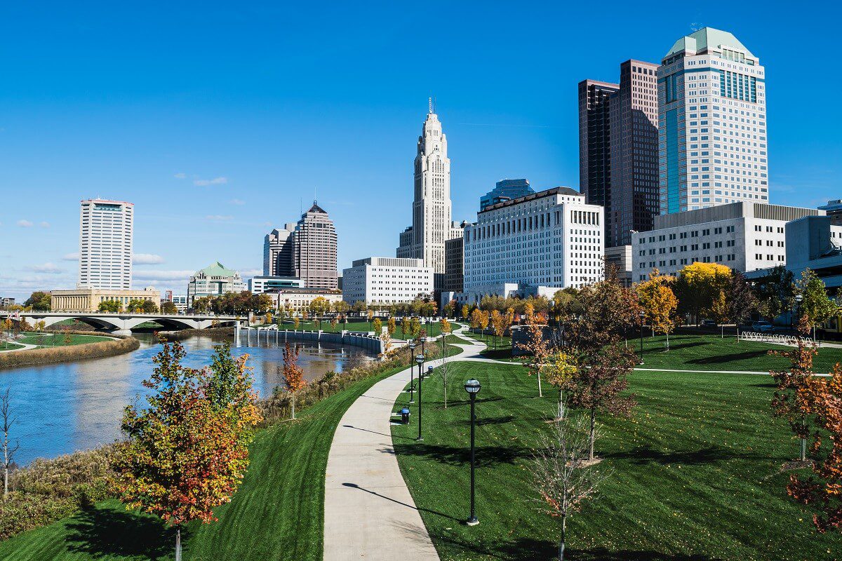 A city skyline with tall buildings stands beside a river and a green park with a winding path and autumn trees under a clear blue sky.