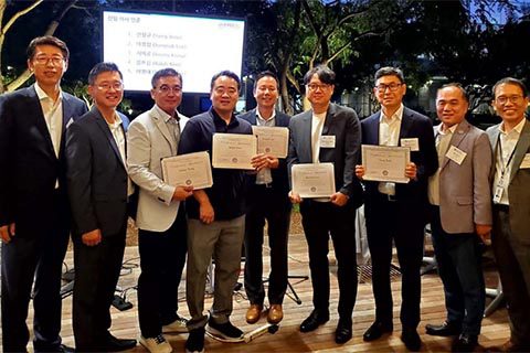 A group of men in business attire stand together outdoors, with several holding certificates and smiling at the camera. A presentation screen is visible in the background.