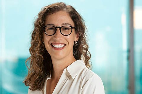 Woman with curly brown hair and glasses, wearing a white blouse, smiling in front of a blurred blue background.