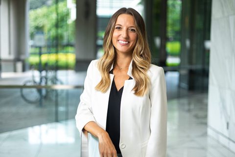 A woman with long, wavy hair wearing a white blazer and black top stands indoors, smiling, in a modern building with glass walls and greenery outside.