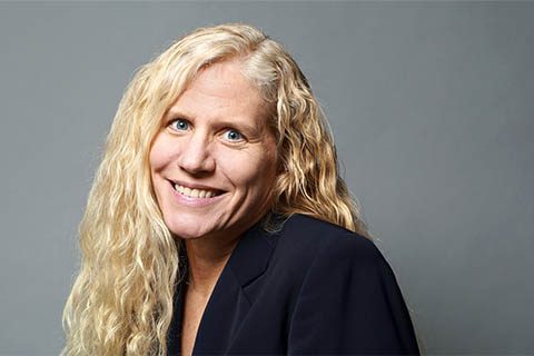 A person with long, wavy blonde hair wearing a dark blazer, smiling at the camera against a plain gray background.