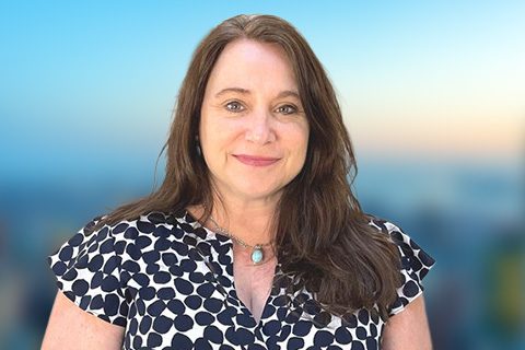 Woman with long brown hair wearing a patterned blouse and necklace, standing in front of a blurred outdoor background with blue sky.