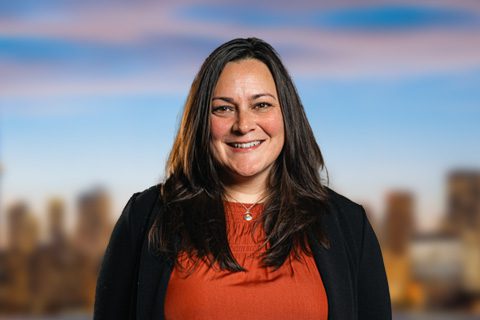 A woman with long brown hair smiles at the camera, wearing an orange top and black jacket, with a blurred cityscape in the background.