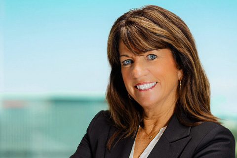 A woman with brown hair wearing a black blazer and white top smiles at the camera with a blurred background.