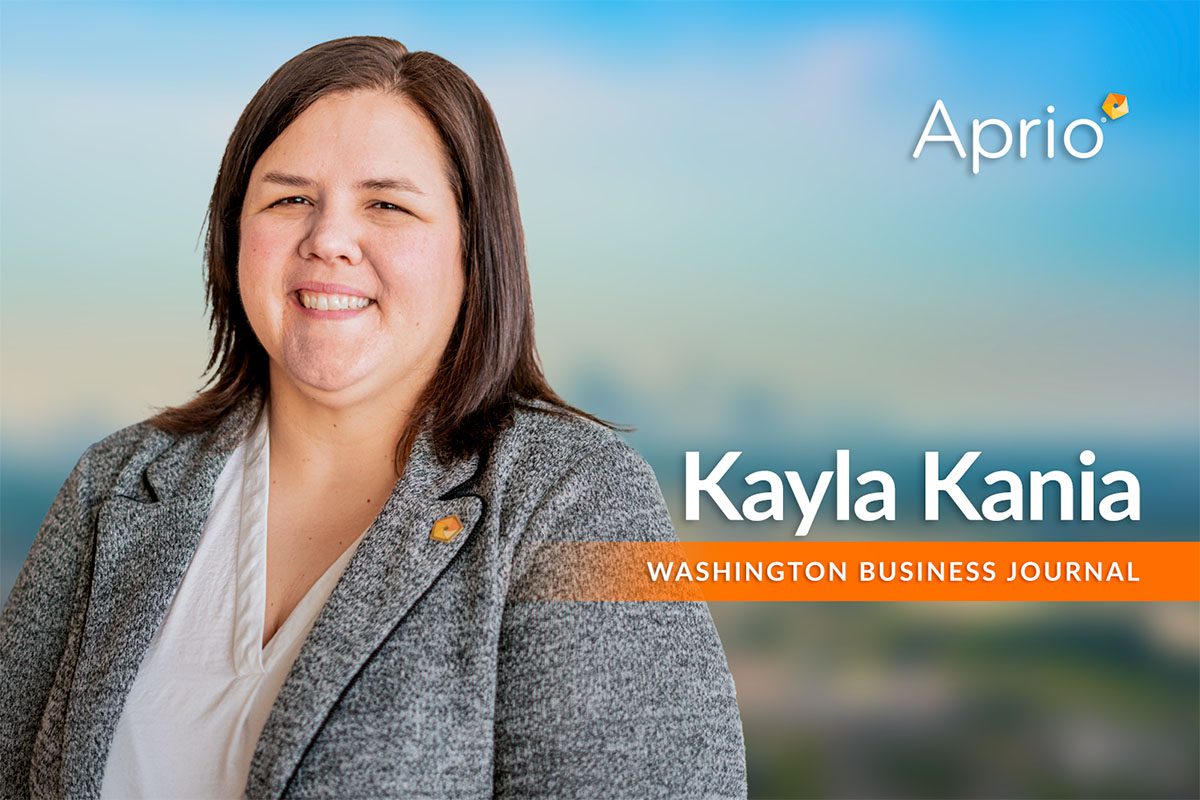 A woman in a gray blazer and white shirt stands smiling in front of a blurred background, with the text Aprio, Kayla Kania, and Washington Business Journal displayed.