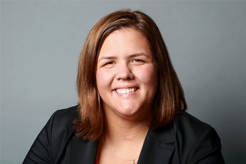 A woman with straight brown hair, wearing a black blazer, smiles at the camera against a plain gray background.