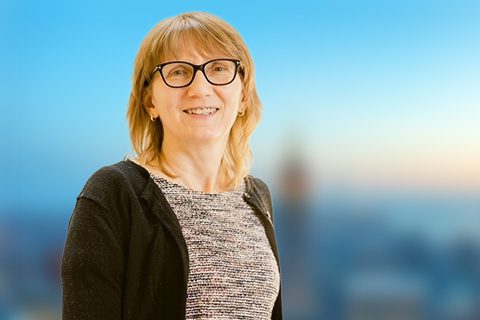 A woman with glasses and shoulder-length light brown hair is smiling, standing in front of a blurred cityscape background.
