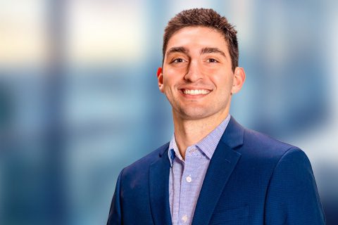 A man wearing a blue suit jacket and light blue shirt smiles while standing in front of a blurred office background.