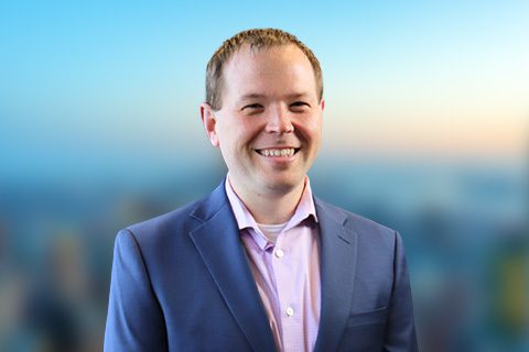 Man in a blue blazer smiles in front of a bright window with a blurred city view.