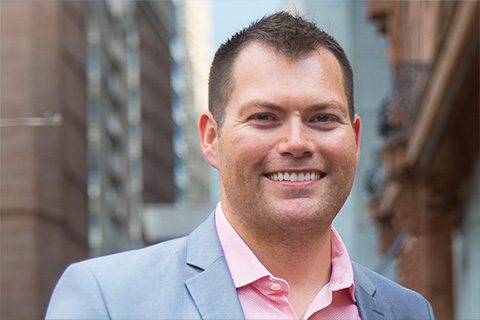 A man in a light gray blazer and pink shirt smiles outdoors in an urban setting with tall buildings in the background.