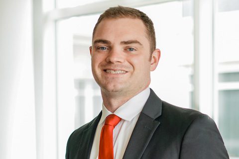 A man in a dark suit, white shirt, and red tie is smiling, standing in front of large windows with natural light coming in.