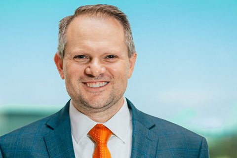 A man in a blue suit, white shirt, and orange tie smiles at the camera with a blurred blue and green background.