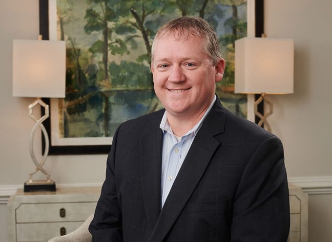 A man in a dark suit and light shirt sits in a room with two lamps and a framed painting of trees in the background.