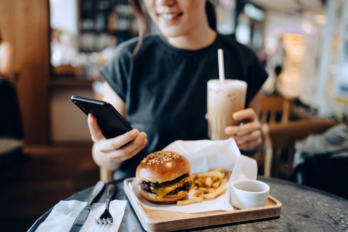 A person sits at a table with a cheeseburger, fries, and a dipping sauce, holding an iced coffee and using a smartphone.
