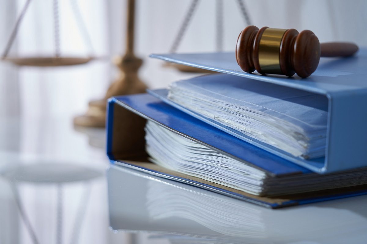 A wooden gavel rests on two blue binders filled with documents, with a blurred scale of justice in the background.