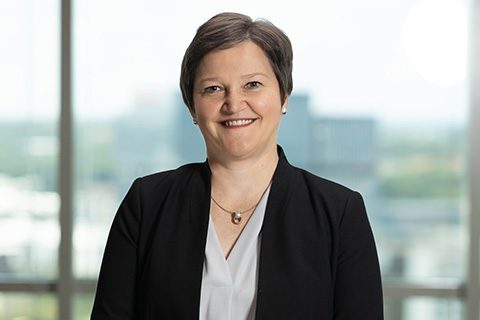 A woman with short brown hair, wearing a black blazer and white blouse, smiles while standing in front of large windows with a blurred cityscape in the background.