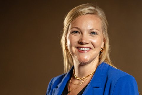 A woman with blonde hair wearing a blue blazer and gold jewelry smiles in front of a plain brown background.
