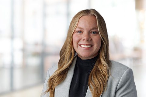 A woman with long blonde hair, wearing a light gray blazer and black top, smiles at the camera with a blurred office background.