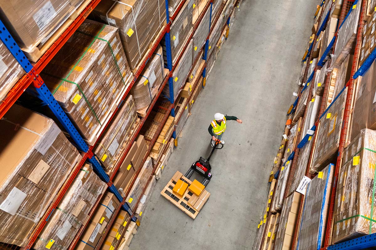 A worker in a safety vest and helmet operates a pallet jack in a warehouse aisle lined with tall shelves of boxed inventory.