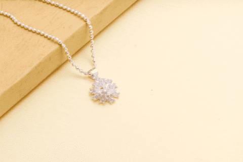 A silver necklace with a snowflake-shaped pendant is displayed on a light yellow surface next to a wooden block.