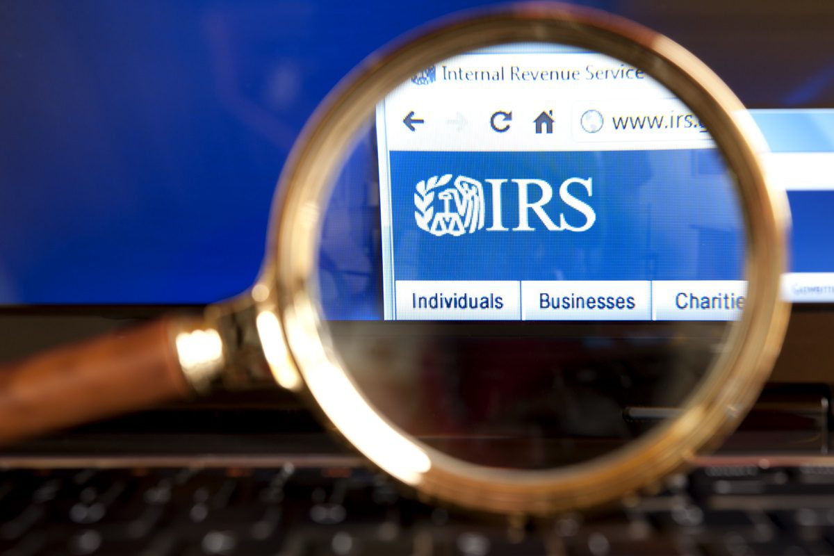 A magnifying glass held over a computer screen highlights the IRS website logo and navigation links for Individuals, Businesses, and Charities.