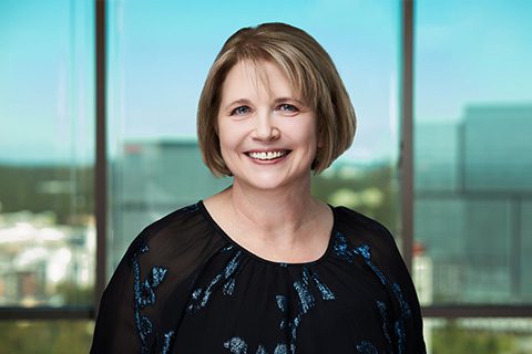 A woman with short blonde hair smiles at the camera, standing indoors in front of large windows showing a cityscape background.
