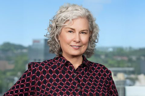 Woman with curly silver hair and a black and red blouse smiles in front of a bright window with a blurred city view.