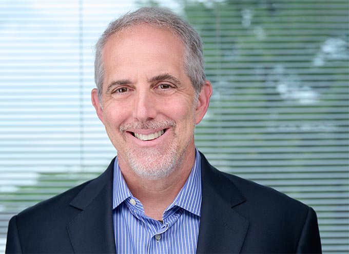 Smiling middle-aged man with short gray hair and beard, wearing a striped blue shirt and dark blazer, standing in front of window blinds.