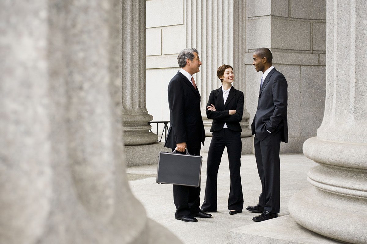 Three business professionals in suits stand talking outside a large stone building with columns; one holds a briefcase.