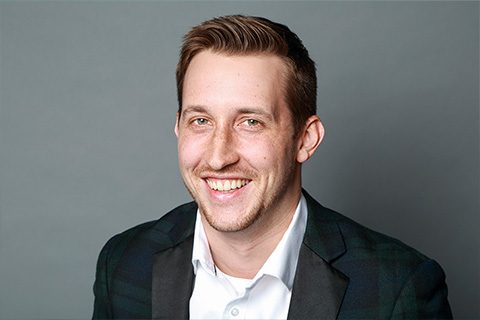 Man with short light brown hair and trimmed beard, wearing a white shirt and dark blazer, smiling in front of a plain gray background.