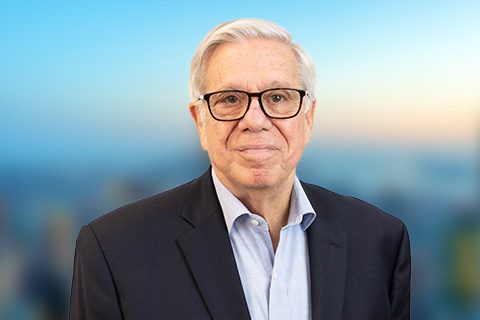 White haired man in glasses and a blazer smiles in front of a bright window with a blurred city view